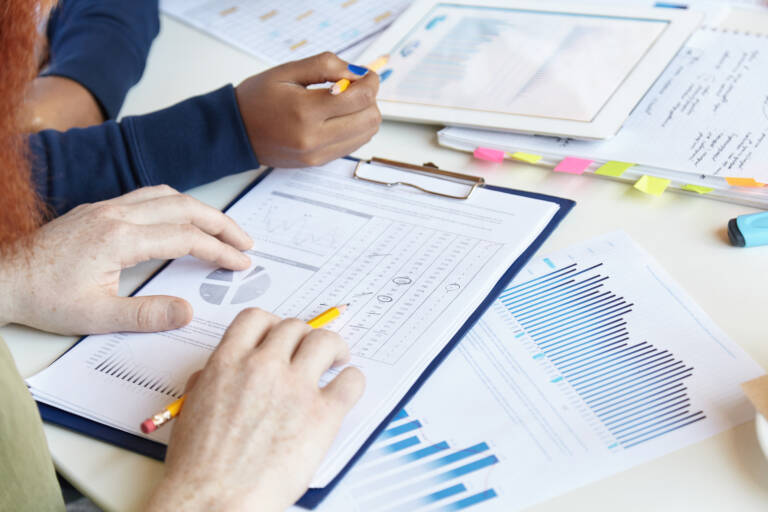 Working in team. Cropped shot of young man holding pencil, studying notes with financial data while sitting at coworking space with his African colleague, discussing business plans and ideas